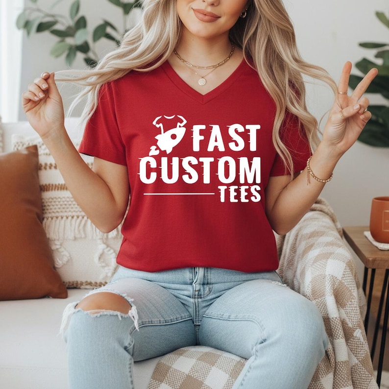 May include: A red V-neck t-shirt with the white text "FAST CUSTOM TEES" and a rocket ship graphic. The shirt is worn by a person wearing light blue jeans.