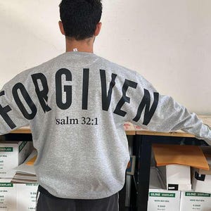 May include: A gray sweatshirt with the word "FORGIVEN" printed in large, black letters across the back. Below the word is the text "salm 32:1". The sweatshirt is a crew neck style.
