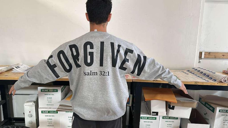 May include: A gray sweatshirt with the word "FORGIVEN" printed in large, black letters across the back. Below the word is the text "salm 32:1". The sweatshirt is a crew neck style.