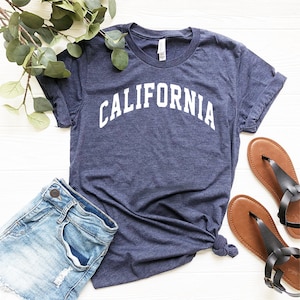 May include: A navy blue t-shirt with the word "CALIFORNIA" printed in white on the front. The shirt is laid flat on a white surface with a pair of brown sandals and a pair of blue denim shorts.