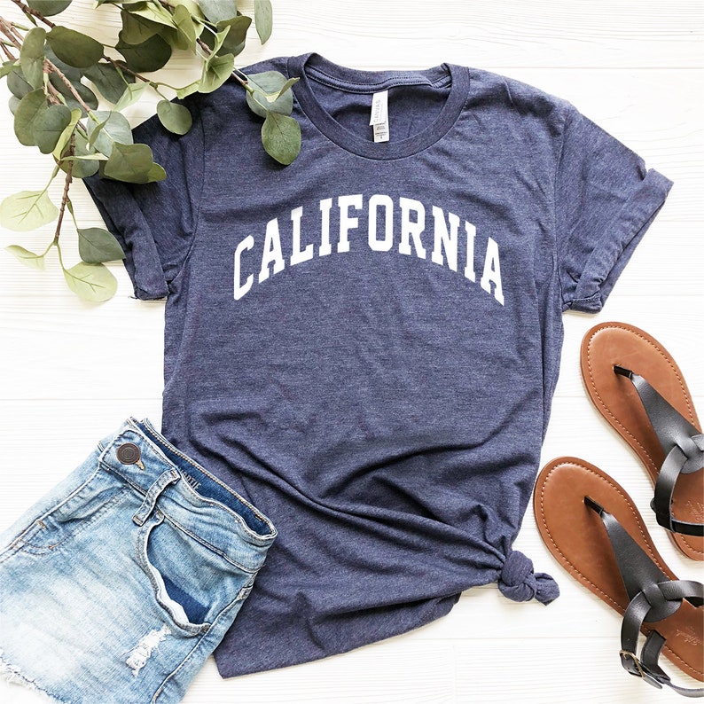 May include: A navy blue t-shirt with the word "CALIFORNIA" printed in white on the front. The shirt is laid flat on a white surface with a pair of brown sandals and a pair of blue denim shorts.