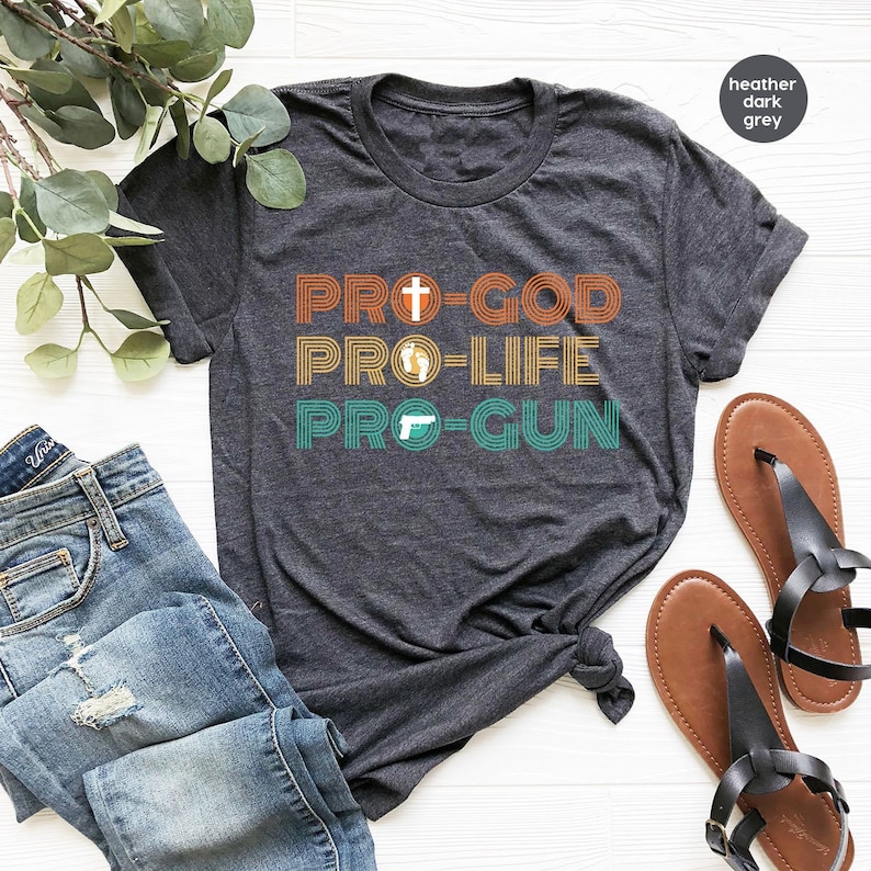 May include: Dark grey t-shirt featuring the text "PRO-GOD PRO-LIFE PRO-GUN" in orange, yellow, and teal. The design includes a cross, footprints, and a handgun. The shirt is shown with blue jeans and sandals.