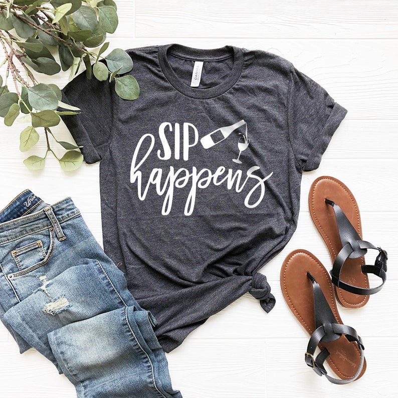 May include: A dark gray t-shirt with the text "SIP happens" in white lettering. The text is accompanied by a white graphic of a wine bottle pouring into a wine glass.