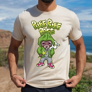 May include: Beige t-shirt featuring a cartoon monkey in a green hoodie, smoking and holding a cannabis leaf. The shirt has the text "PUFF PUFF PASS" in green bubble letters. The monkey is wearing gray pants and pink shoes.