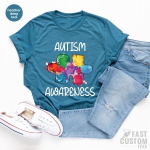 May include: A teal blue t-shirt with the words "Autism Awareness" in white text. A colorful puzzle piece with rainbow colors is in the center of the shirt.