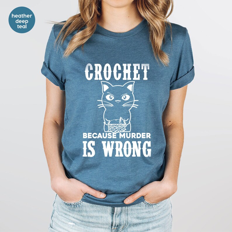 Op de afbeelding: Een heidekleurig diepblauw t-shirt met een witte afbeelding van een breiende kat en de tekst "Haakwerk omdat moord verkeerd is."