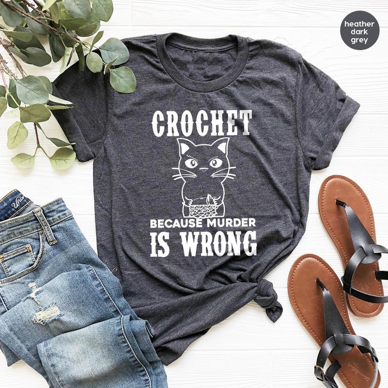 Op de afbeelding: Een donkergrijs t-shirt met een witte afbeelding van een breiende kat en de tekst "CROCHET BECAUSE MURDER IS WRONG".