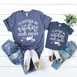 Pode incluir: Duas t-shirts azul-marinho mesclado com o texto branco I'D RATHER BE watching CRIME SHOWS sobre um gr&aacute;fico de salpicos de tinta branca. As t-shirts s&atilde;o combinadas com duas cal&ccedil;as de ganga azuis e dois pares de t&eacute;nis brancos.