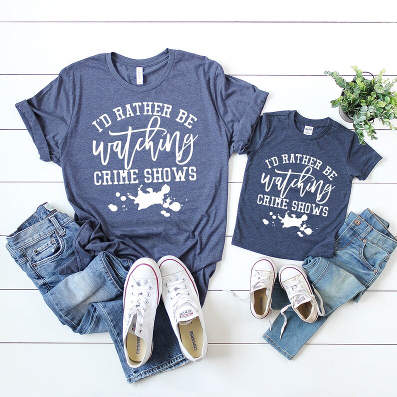 Pode incluir: Duas t-shirts azul-marinho mesclado com o texto branco I'D RATHER BE watching CRIME SHOWS sobre um gr&aacute;fico de salpicos de tinta branca. As t-shirts s&atilde;o combinadas com duas cal&ccedil;as de ganga azuis e dois pares de t&eacute;nis brancos.