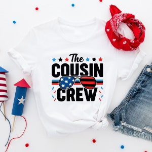 May include: White t-shirt with a red, white, and blue American flag design. The text "The Cousin Crew" is printed in black with a pair of sunglasses with an American flag design.