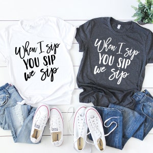 以下が含まれることがあります： 黒い文字で「When I sip YOU SIP we sip」と書かれた白いTシャツが2枚。1枚は白、もう1枚は濃いグレー。Tシャツは白い木製の表面に置かれ、白いスニーカーが2足と青いジーンズが2本置かれている。