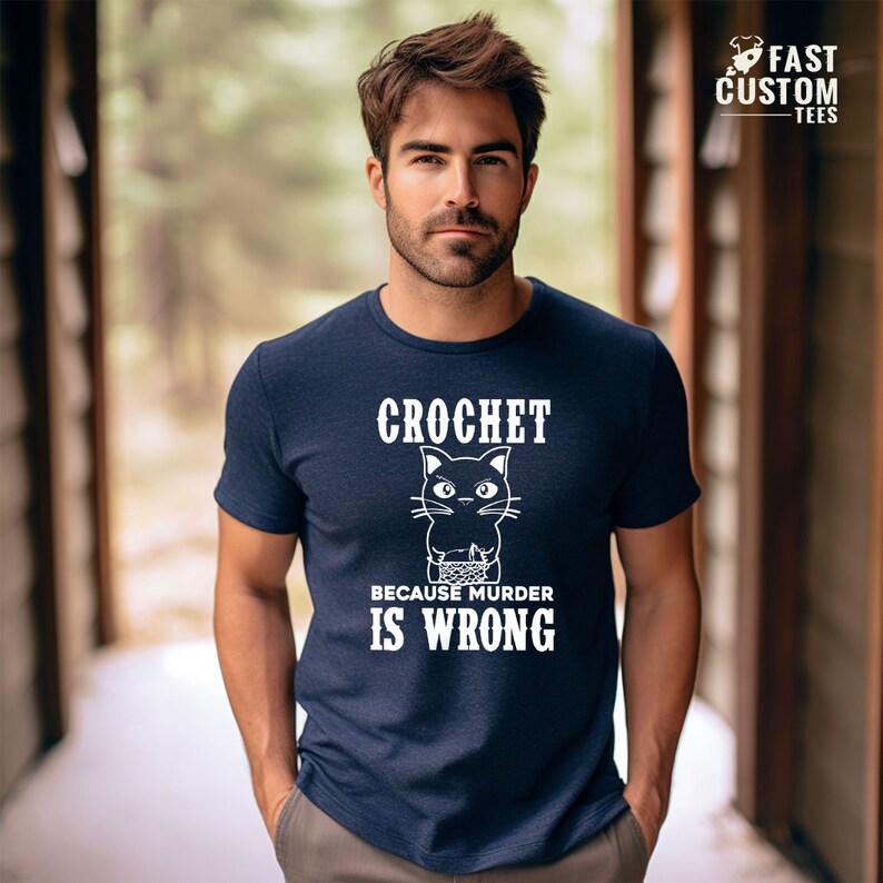 Op de afbeelding: Een marineblauw t-shirt met een witte afbeelding van een zwarte kat die haakt met de tekst "Crochet because murder is wrong".