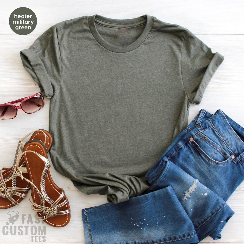 May include: A plain, heather green t-shirt with rolled up sleeves. The t-shirt is laid flat on a white wooden surface with a pair of brown sandals, blue jeans, and sunglasses.