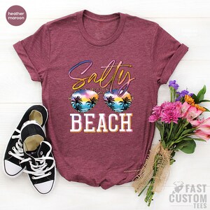 May include: A heather maroon t-shirt with the text "Salty Beach" in colorful letters. The text is overlaid with a pair of sunglasses with a sunset and palm trees reflected in the lenses.