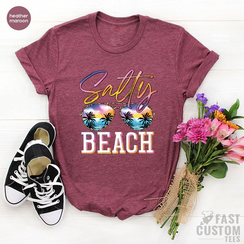 May include: A heather maroon t-shirt with the text "Salty Beach" in colorful letters. The text is overlaid with a pair of sunglasses with a sunset and palm trees reflected in the lenses.