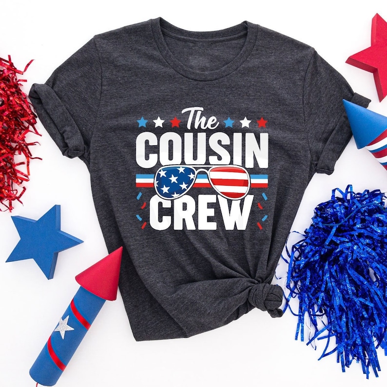 May include: A dark gray t-shirt with the text "The Cousin Crew" in white. The text is surrounded by red, white, and blue stars and confetti. The shirt also features a pair of sunglasses with an American flag design.