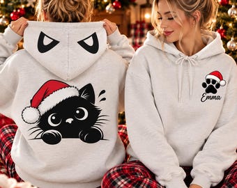 May include: A light gray hooded sweatshirt featuring a black cat graphic wearing a red and white Santa hat on the back, with matching cat ear designs on the hood. The front displays a black paw print with a Santa hat and the name Emma.