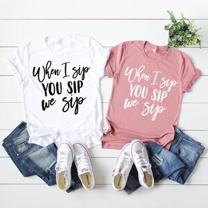 以下が含まれることがあります： 黒い文字で「When I sip YOU SIP we sip」と書かれた白いTシャツが2枚。1枚は白、もう1枚はピンク。白い木製の表面に、白いスニーカー2足と青いジーンズ2本と一緒に置かれている。