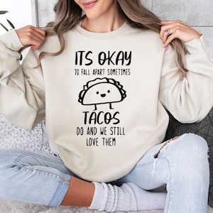 May include: A white sweatshirt with a black graphic that says "It's okay to fall apart sometimes tacos do and we still love them". The graphic features a cartoon taco with a smiling face.