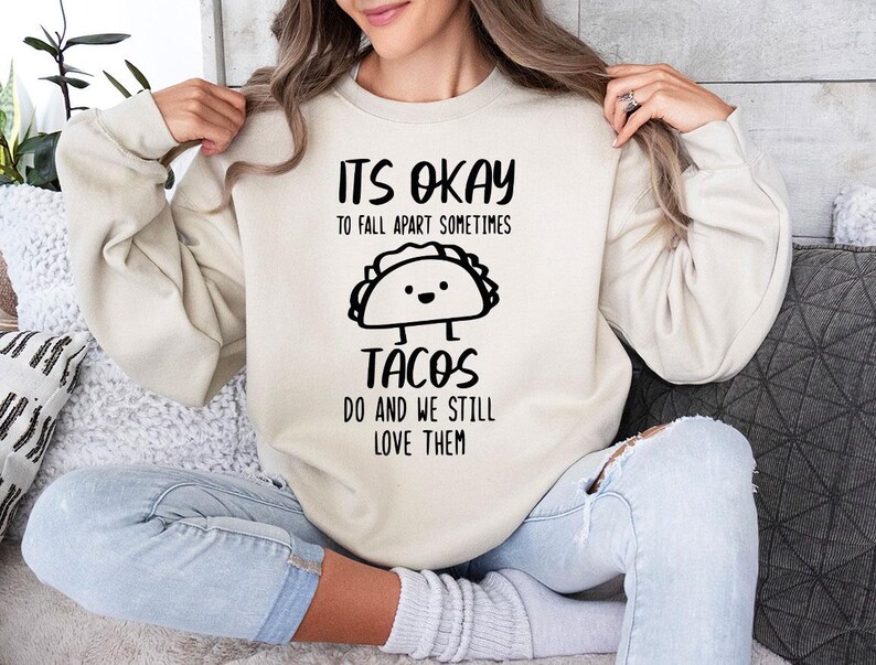May include: A white sweatshirt with a black graphic that says "It's okay to fall apart sometimes tacos do and we still love them". The graphic features a cartoon taco with a smiling face.