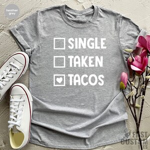 May include: Heather grey t-shirt with a white graphic that says "Single, Taken, Tacos" with a heart in the box next to "Tacos".