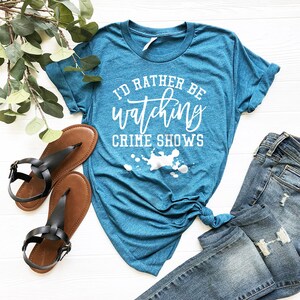 Pode incluir: Uma t-shirt de manga curta azul-petr&oacute;leo com o texto branco I'D RATHER BE watching CRIME SHOWS sobre um gr&aacute;fico de salpicos de tinta branca. A t-shirt &eacute; combinada com sand&aacute;lias pretas e cal&ccedil;as de ganga azuis com detalhes desgastados.