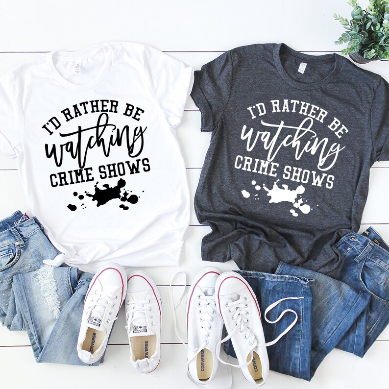 Pode incluir: Duas t-shirts de manga curta, uma branca com texto preto e uma cinzento-escura com texto branco, ambas com a frase I'D RATHER BE watching CRIME SHOWS e um gr&aacute;fico de salpicos de tinta. Duas cal&ccedil;as de ganga azuis e dois pares de t&eacute;nis brancos.