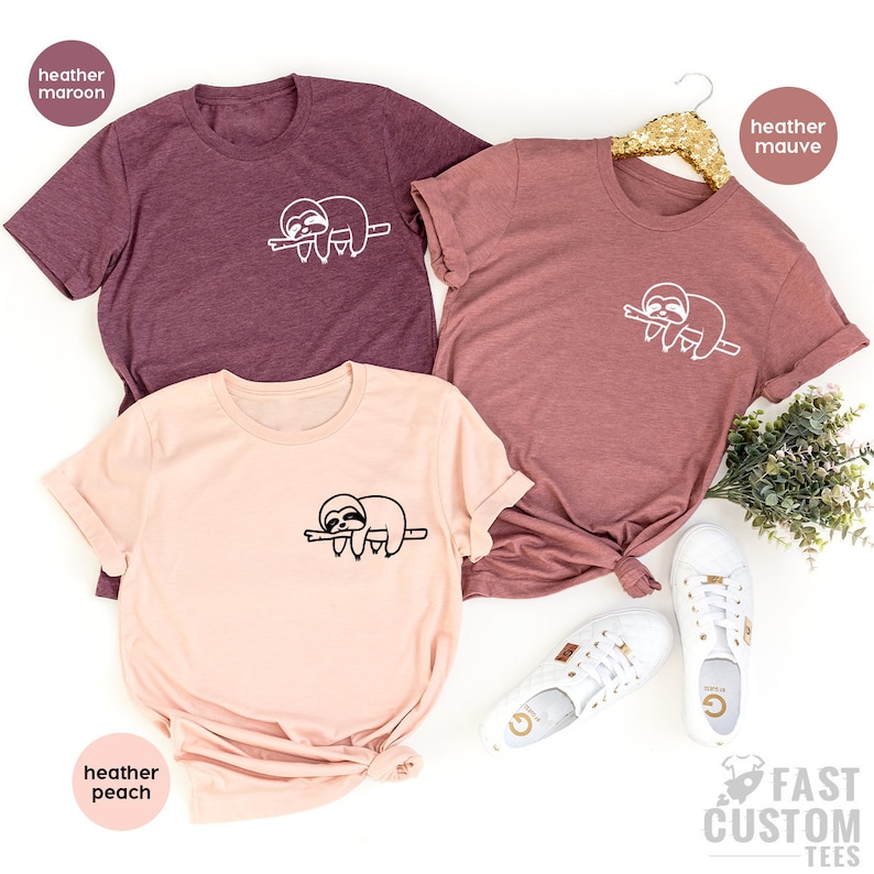 K&ouml;nnte beinhalten: Drei T-Shirts mit einem Faultier-Motiv. Die Shirts sind in den Farben Heidelbeer-Marone, Heidelbeer-Lila und Heidelbeer-Pfirsich gehalten. Das Faultier ist schlafend auf einem Ast dargestellt.