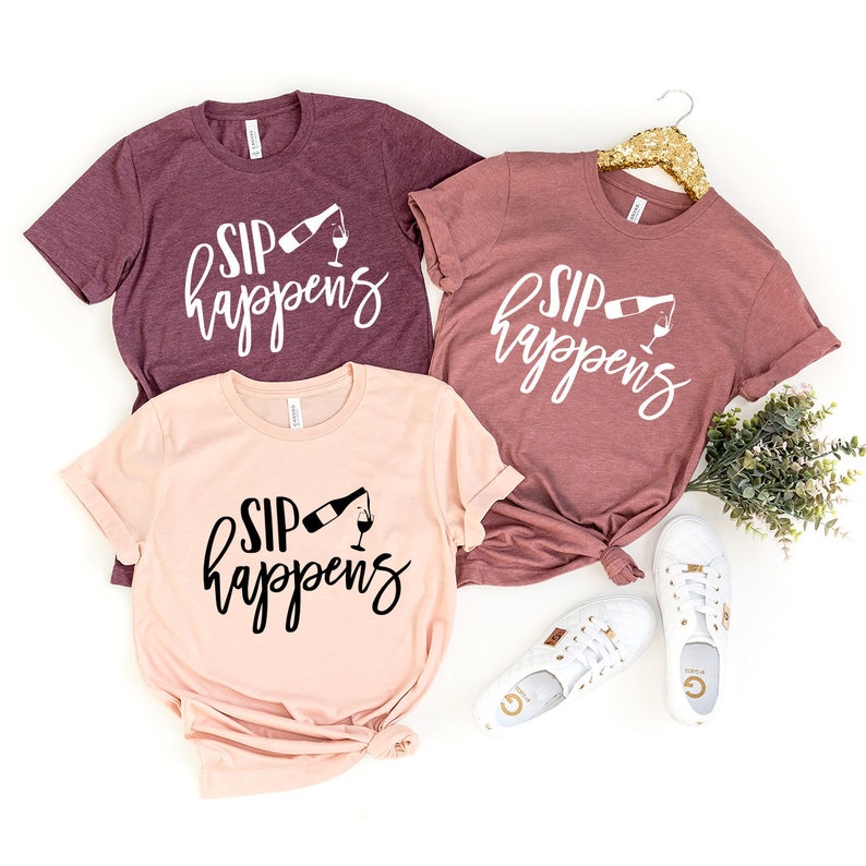 May include: Three t-shirts with the text "Sip Happens" and a graphic of a wine bottle pouring into a glass. The shirts are in different colors: maroon, light pink, and a darker shade of pink.