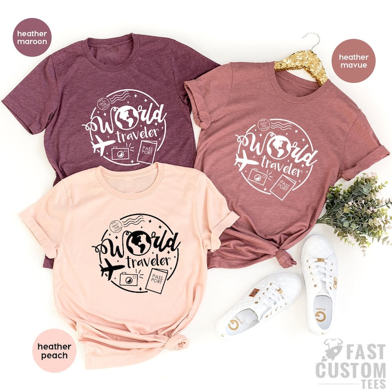 May include: Three t-shirts with the text "World Traveler" in a circle with a globe, airplane, camera, and passport. The shirts are in heather maroon, heather peach, and heather mauve.
