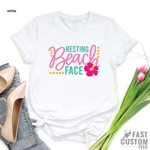 May include: White t-shirt with the text "Resting Beach Face" in teal and pink lettering with a pink hibiscus flower.