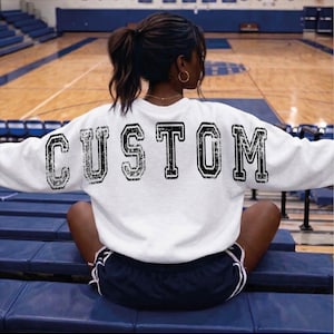 May include: A white sweatshirt with the word "CUSTOM" printed across the back in a distressed, black, collegiate-style font. The sweatshirt is paired with navy blue athletic shorts with white stripes.