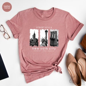 May include: Heather mauve t-shirt with a graphic print of the Empire State Building, the Statue of Liberty, and the Brooklyn Bridge. The text "Street Style" and "New York City" is printed on the shirt.