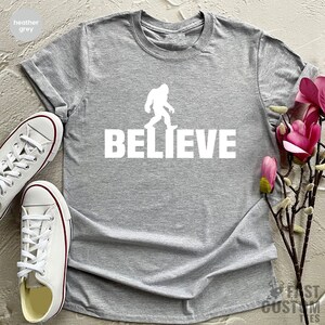 May include: Heather grey t-shirt with a white silhouette of Bigfoot walking and the word "BELIEVE" printed below.