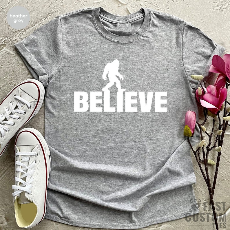 May include: Heather grey t-shirt with a white silhouette of Bigfoot walking and the word "BELIEVE" printed below.