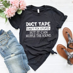 May include: Black t-shirt with white text that reads "Duct Tape Can't Fix Stupid But It Can Muffle The Sound". The shirt is laid out on a white surface with a pair of blue jeans and brown sandals.