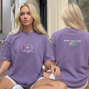 May include: A purple short-sleeve t-shirt featuring a graphic of a battery icon at 5% with the text "still running" on the front, and the text "power save mode on" with a green battery icon on the back of the garment.