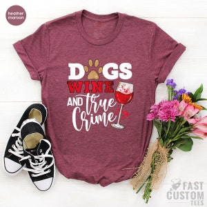 May include: Heather maroon t-shirt with the text "Dogs, Wine, and True Crime" in white and red. A paw print and a wine glass with red wine are also featured. Black canvas sneakers and a bouquet of flowers are in the image.