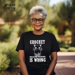Op de afbeelding: Een vrouw draagt een zwart T-shirt met een witte afbeelding van een zwarte kat die haakt. De tekst op het shirt luidt "Crochet omdat moord verkeerd is."