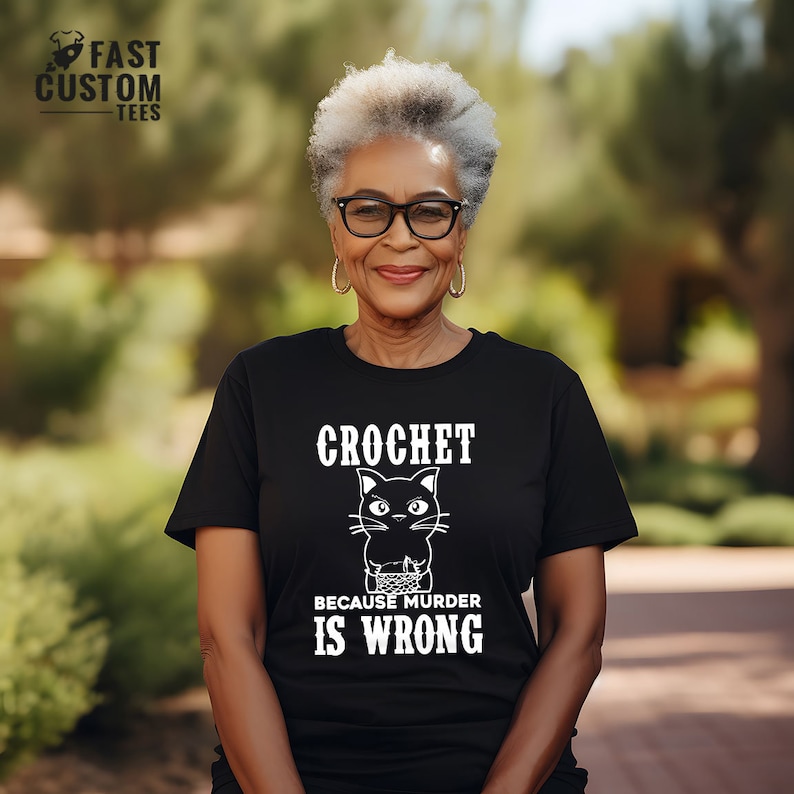 Op de afbeelding: Een vrouw draagt een zwart T-shirt met een witte afbeelding van een zwarte kat die haakt. De tekst op het shirt luidt "Crochet omdat moord verkeerd is."