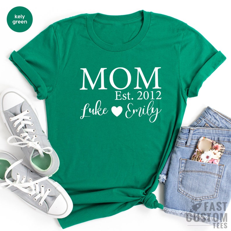 May include: A kelly green t-shirt with the text "MOM Est. 2012 Luke Emily" in white. The text "MOM" is in a large, bold font, while the rest of the text is in a smaller, cursive font. A heart symbol is between the names Luke and Emily.