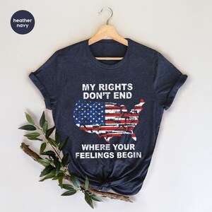 May include: Navy blue t-shirt featuring the text "MY RIGHTS DON'T END WHERE YOUR FEELINGS BEGIN" above a distressed American flag design in the shape of the United States. The shirt is displayed on a wooden hanger.