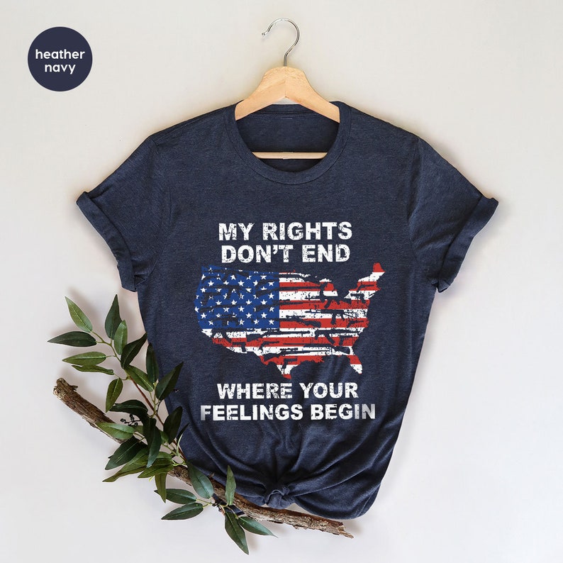 May include: Navy blue t-shirt featuring the text "MY RIGHTS DON'T END WHERE YOUR FEELINGS BEGIN" above a distressed American flag design in the shape of the United States. The shirt is displayed on a wooden hanger.