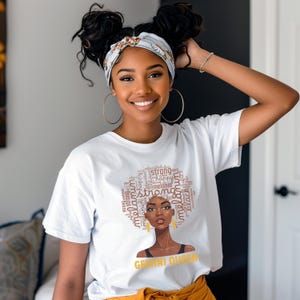 May include: A white t-shirt featuring a graphic of a woman's face with the words "strong", "melanin", and "happy" in a circular design around her hair. The text "Gemini Queen" is below the graphic. The woman is wearing gold hoop earrings.