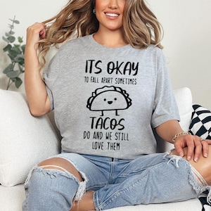 May include: A gray t-shirt with black text that reads "It's okay to fall apart sometimes tacos do and we still love them". The shirt features a cartoon taco with a smiling face.