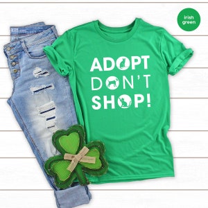 May include: A green t-shirt with the text "ADOPT DON'T SHOP!" in white. The text is surrounded by circles with a cat and dog silhouette in each circle. The shirt is laid out on a white wooden surface with a pair of blue jeans and a green shamrock.