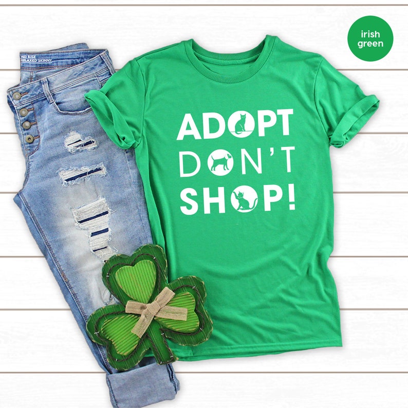 May include: A green t-shirt with the text "ADOPT DON'T SHOP!" in white. The text is surrounded by circles with a cat and dog silhouette in each circle. The shirt is laid out on a white wooden surface with a pair of blue jeans and a green shamrock.