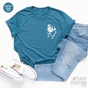 May include: A heather deep teal t-shirt with a white pug graphic on the front pocket. The shirt is laid flat on a white surface with a pair of white trainers and light blue jeans.