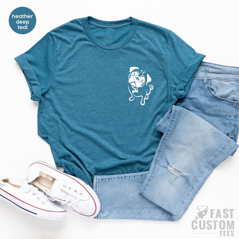May include: A heather deep teal t-shirt with a white pug graphic on the front pocket. The shirt is laid flat on a white surface with a pair of white trainers and light blue jeans.