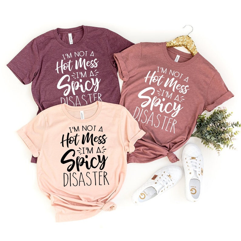 May include: Three t-shirts with the text "I'm not a hot mess, I'm a spicy disaster" in black and white lettering. The shirts are in different colors: maroon, pink, and a lighter shade of maroon.